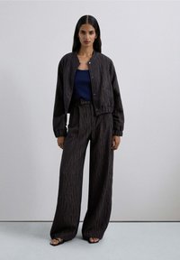 Mujer vistiendo una chaqueta oscura de patrón suelto y pantalones de pierna ancha con un top azul marino y sandalias negras, de pie contra un fondo claro y liso.