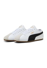 Baskets blanches avec accents en daim crème et noir. Elles présentent des lacets plats, un bout arrondi et une semelle en caoutchouc. Détail du logo doré sur le côté.