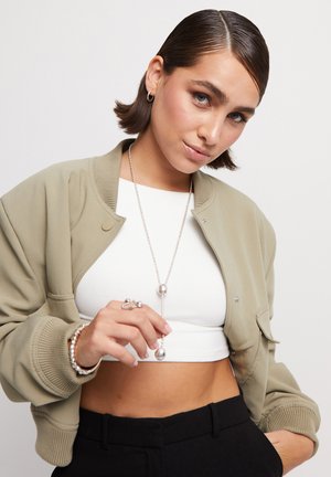 Chaqueta bomber verde oliva, con puños acanalados, llevada sobre una blusa corta blanca. Se pueden ver joyas plateadas y un pantalón negro de tiro alto.