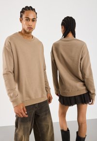To personer med flettede hår har matchende beige hettegensere; den ene står vendt fremover, den andre viser ryggen, kombinert med mørke bukser.