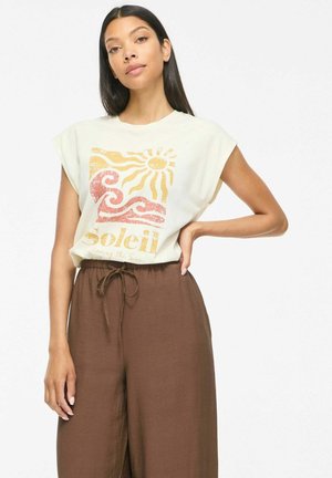 Mujer con cabello largo y oscuro que lleva una camiseta blanca sin mangas con diseño de sol y olas y el texto "Soleil", combinada con pantalones marrones sueltos con cordón.