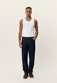 Λευκό αμάνικο μπλουζάκι με μαύρο λογότυπο, σε συνδυασμό με σκούρα μπλε τζιν. Το μοντέλο φοράει καφέ παπούτσια, με τα χέρια στις τσέπες. Απλός, καθημερινός σχεδιασμός.