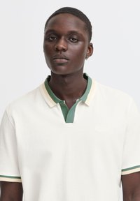Polo en algodón color crema con cuello verde y acentos a rayas. Presenta un escote en V y mangas cortas. Logo sutil bordado en el pecho.