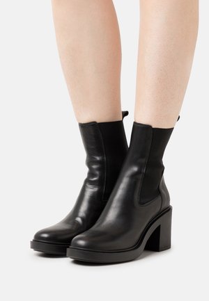 Bottes cheville en cuir noir avec un talon épais, des panneaux latéraux élastiques et une finition lisse. Le design présente une esthétique minimaliste.