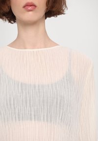 Gros plan d'une personne portant un haut blanc transparent texturé avec une sous-couche grise visible et des lèvres roses brillantes.