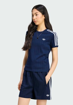 Young woman standing wearing navy blue Adidas sportswear with white stripes, including a short-sleeve shirt and matching shorts.