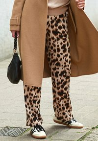 Luipaardprint broek in beige en zwart, gecombineerd met een camelkleurige jas. Gestyled met een zwarte tas en witte sneakers met zwarte accenten.