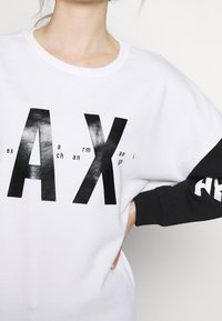 Weißes Sweatshirt mit auffälligen schwarzen grafischen Buchstaben "AX" auf der Vorderseite, mit langen schwarzen Ärmeln und geripptem Halsausschnitt.