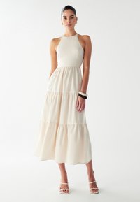 Nainen, jolla on yllään hihaton beige kerroksellinen midimekko ja valkoiset nauhakengät, asusteinaan kultaketjuiset korvakorut ja kaksi suurta rannetta.