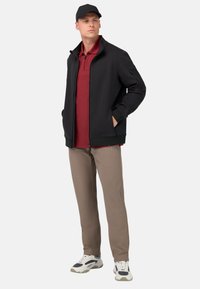 Črna jakna na zadrgo, maroon polo majica, bež hlače in bele teniske. Model pozira z rokami v žepih jakne, na glavi nosi črno kapo.