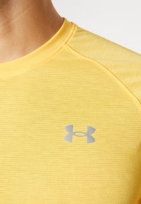 Žluté sportovní tričko s texturovanou tkaninou. Má kulatý výstřih a stříbrné logo na levé části hrudi.