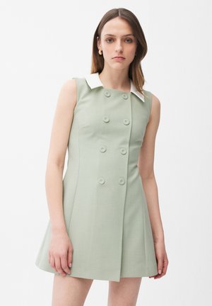 Giovane donna che indossa un vestito verde chiaro senza maniche con colletto bianco, in piedi contro uno sfondo bianco uniforme.