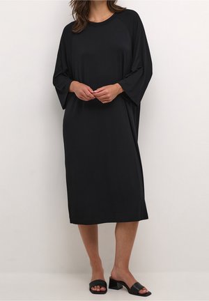 Abito nero al ginocchio realizzato in tessuto morbido, con vestibilità ampia, maniche raglan e scollo rotondo. Indossato con sandali neri a ciabatta.