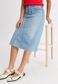 Jupe midi en denim, lavage bleu clair, ourlet effiloché, coutures avant pour la structure. Associée à des chaussures à talons blancs avec de grandes boucles noires.