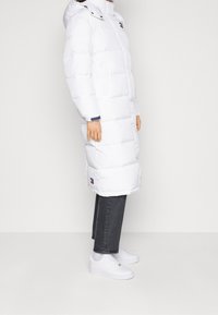 Vit puffjacka med huva, med dragkedjestängning, sidofickor och horisontellt quiltat mönster. I kombination med mörka jeans och vita sneakers.