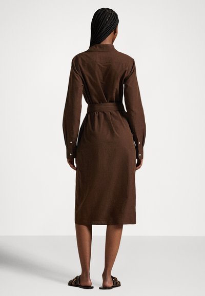 Woman with braided hair wearing a brown long-sleeve belted midi dress and flat sandals, standing facing away on a plain background.