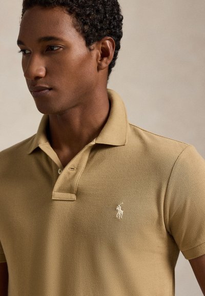 Beige pikétröja i strukturerat tyg med tvåknappslinning och broderad logotyp på bröstet. Har klassisk krage och korta ärmar.