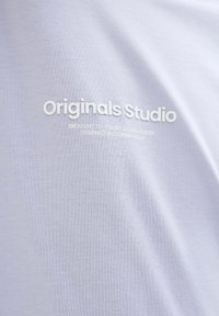 Šviesiai violetinė medvilninė marškinėliai su iškilniu baltu užrašu: "Originals Studio" ir papildomu tekstu apačioje, lygus tekstūras ir kasdienis dizainas.