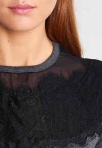 Blusa nera in pizzo con un top trasparente e colletto a costine grigio. I dettagli in pizzo testurizzato sul petto aggiungono un interesse visivo.