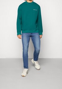 Modrozelená mikina s kulatým výstřihem a žebrovanými manžetami, doplněná modrými džínami a bílými teniskami. Minimalistické logo na hrudi.