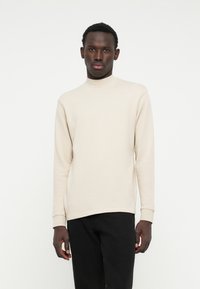 Felpa beige a maniche lunghe con collo alto in morbido tessuto, con polsini a costine, abbinata a jeans neri skinny. Design semplice e moderno.