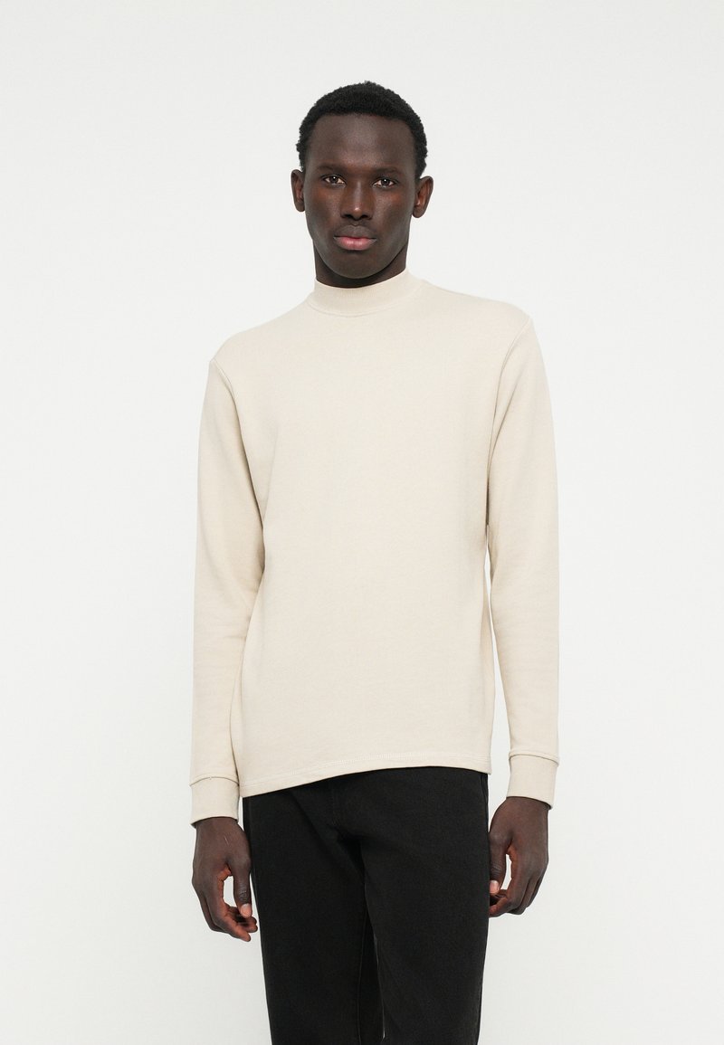 Felpa beige a maniche lunghe con collo alto in morbido tessuto, con polsini a costine, abbinata a jeans neri skinny. Design semplice e moderno.