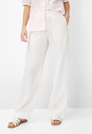 Person iført hvide vidde bukser, en lyserød blomsterprint skjorte med knapper og hvide slip-on sandaler med lysebrune såler.