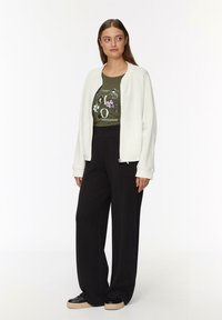 Witte getextureerde bomberjack over een groene graphic T-shirt, gecombineerd met losse zwarte broeken en zwarte platform sneakers.