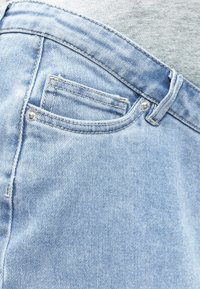 Prim-plan cu buzunarul blugilor din denim albastru deschis, cu cusături vizibile, nituri și cataramă pentru curea, pe un fundal din material gri.