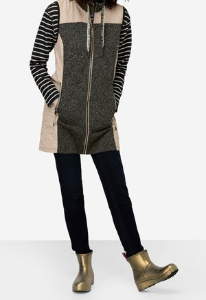 Personne portant un gilet long beige et gris zippé sur une chemise à manches longues rayée noir et blanc, un pantalon noir et des bottines dorées.