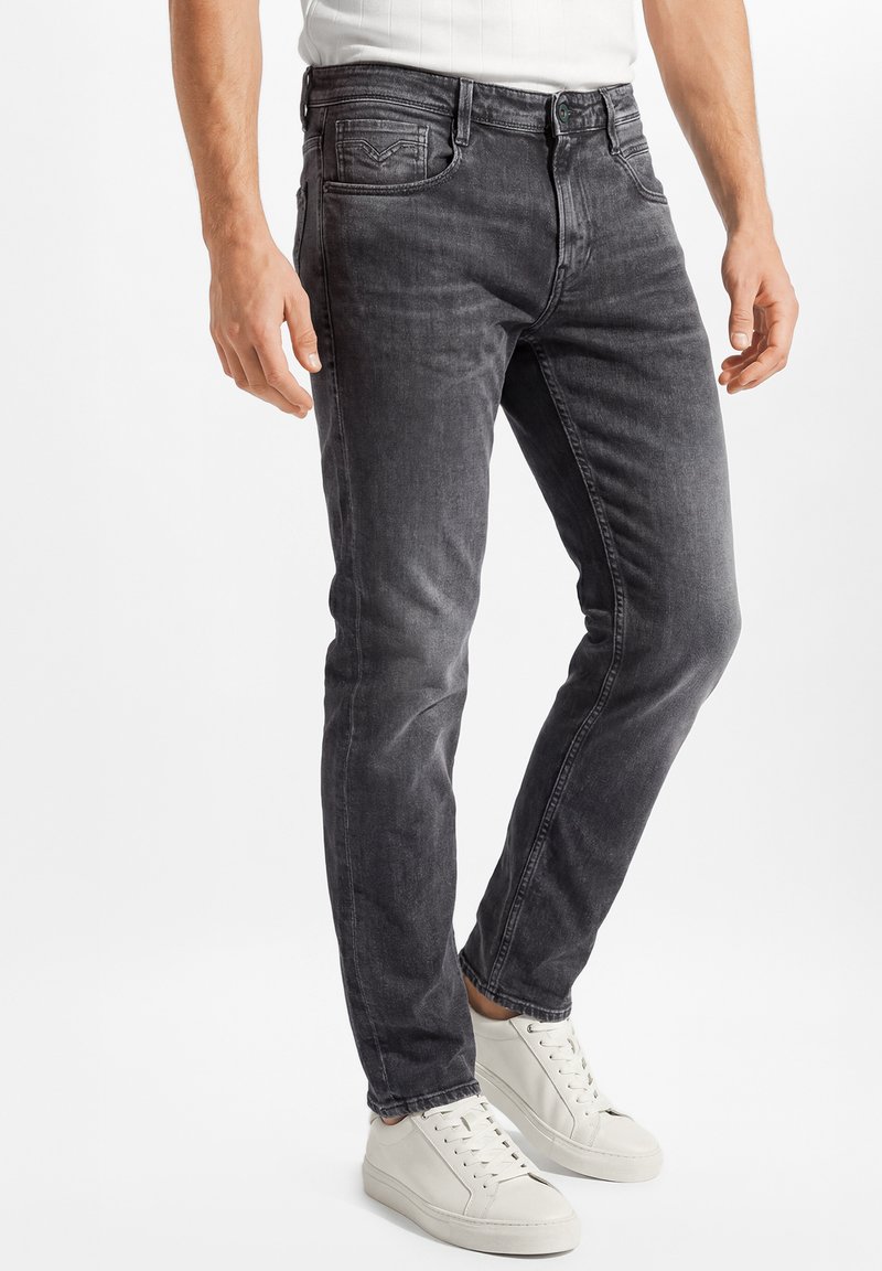 Mann trägt schmal geschnittene, ausgewaschene schwarze Jeans und weiße Sneakers, steht mit locker an den Seiten hängenden Händen vor einfarbigem Hintergrund.