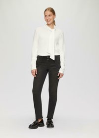 Witte lange mouwen top met een nekstrik, gedragen met zwarte slim-fit jeans en zwarte dikke loafers, op een lichtgrijze achtergrond.