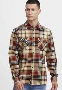 Camisa de franela a cuadros en colores rojo, amarillo, marrón y negro; cuenta con dos bolsillos en el pecho, cuello abotonado y un dobladillo recto.