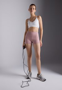 Sujetador deportivo blanco con tirantes finos, pantalones cortos de tiro alto rosas y zapatillas de running plateadas. Sostiene una cuerda con una mano, frente a un fondo gris.