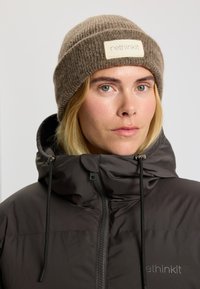 Brun strikket beanie med en foldet kant og et rektangulært mærkat foran, sammen med en mørk, lynlåset pufferjakke.