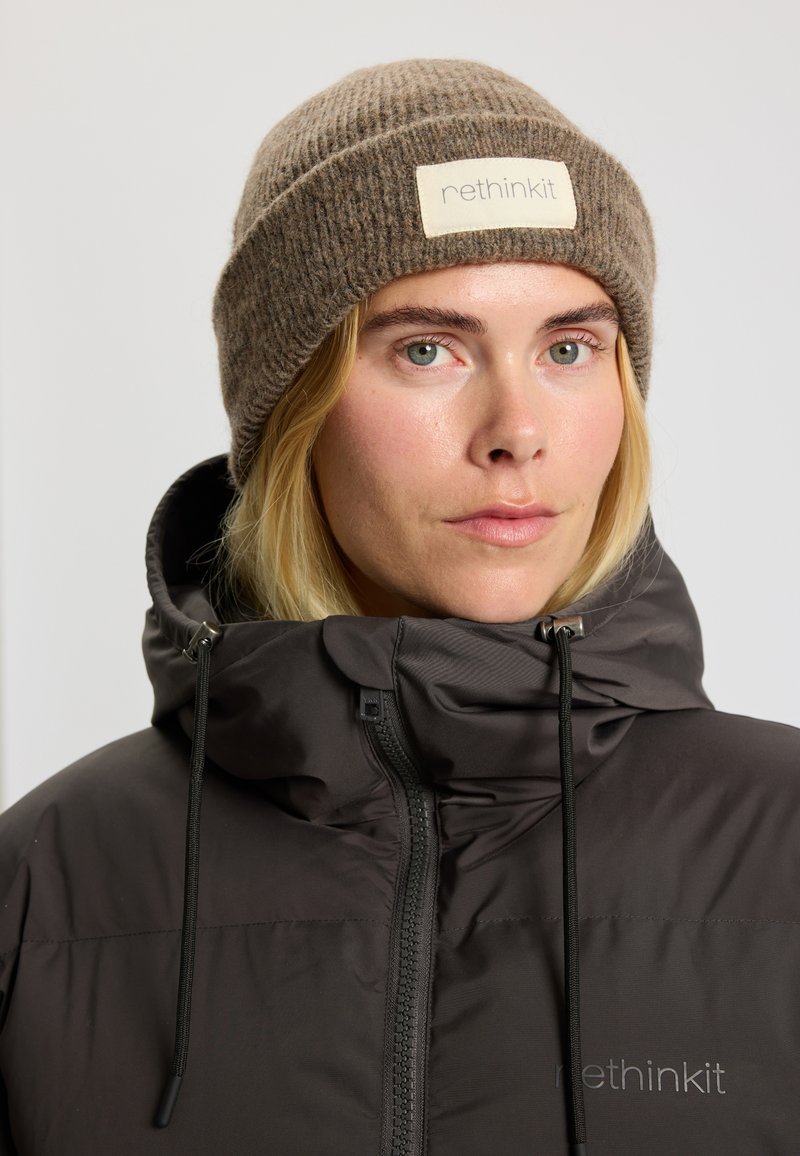 Brun strikket beanie med en foldet kant og et rektangulært mærkat foran, sammen med en mørk, lynlåset pufferjakke.