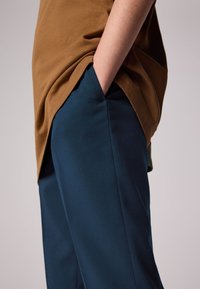 Ruskea t-paita laskeutuu tummansinisille, sileäpintaisille housuille. Housuissa on sivutaskut ja räätälöity istuvuus, siisteillä linjoilla.