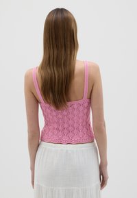Crop top rosa lavorato a maglia con orlo a scallop e motivo in pizzo. Presenta spalline sottili e una finitura testurizzata, abbinato a una gonna bianca fluida.
