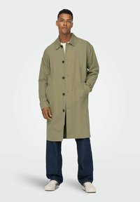 Manteau long vert olive avec un devant boutonné, un large col et des poches latérales, associé à un jean foncé et des baskets blanches. Texture de tissu lisse.