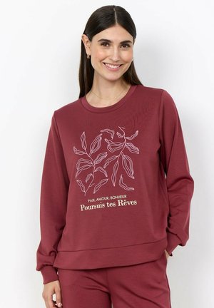 Lachende vrouw met lang donker haar draagt een wijnrood sweatshirt- en broekenset met witte plantillustraties en Franse tekst aan de voorkant.