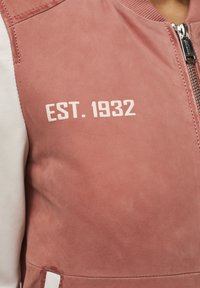 Różowa zamszowa kurtka bombowa z białymi skórzanymi rękawami, z zamkiem błyskawicznym i napisem "EST. 1932" wydrukowanym na klatce piersiowej.
