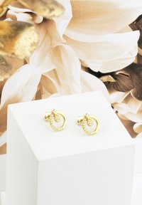 Orecchini d'oro con un design a spirale e un'accento di pietra chiara, presentati su uno sfondo chiaro e mostrati su un piedistallo bianco.