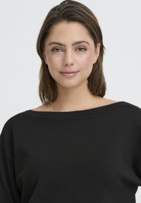 Mladá žena s hnedými vlasmi po pleciach, oblečená v čiernom svetri bez ramienok, jemne sa usmievajúca na jednoduchom svetlom pozadí.