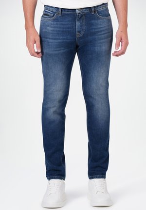 Man wearing slim-fit blue jeans and white sneakers, standing against a plain light background.