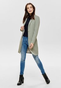 Cardigan lungo verde chiaro con colletto a bavero, chiusura con un bottone e maniche lunghe, abbinato a jeans blu aderenti e stivaletti neri alla caviglia.