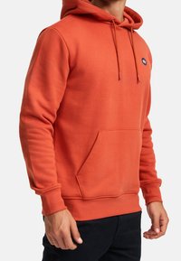 Orange fleece-tröja med kengurufickor, justerbara dragsnören, ribbade ärmmuddar och en liten logotyp på vänster bröst.
