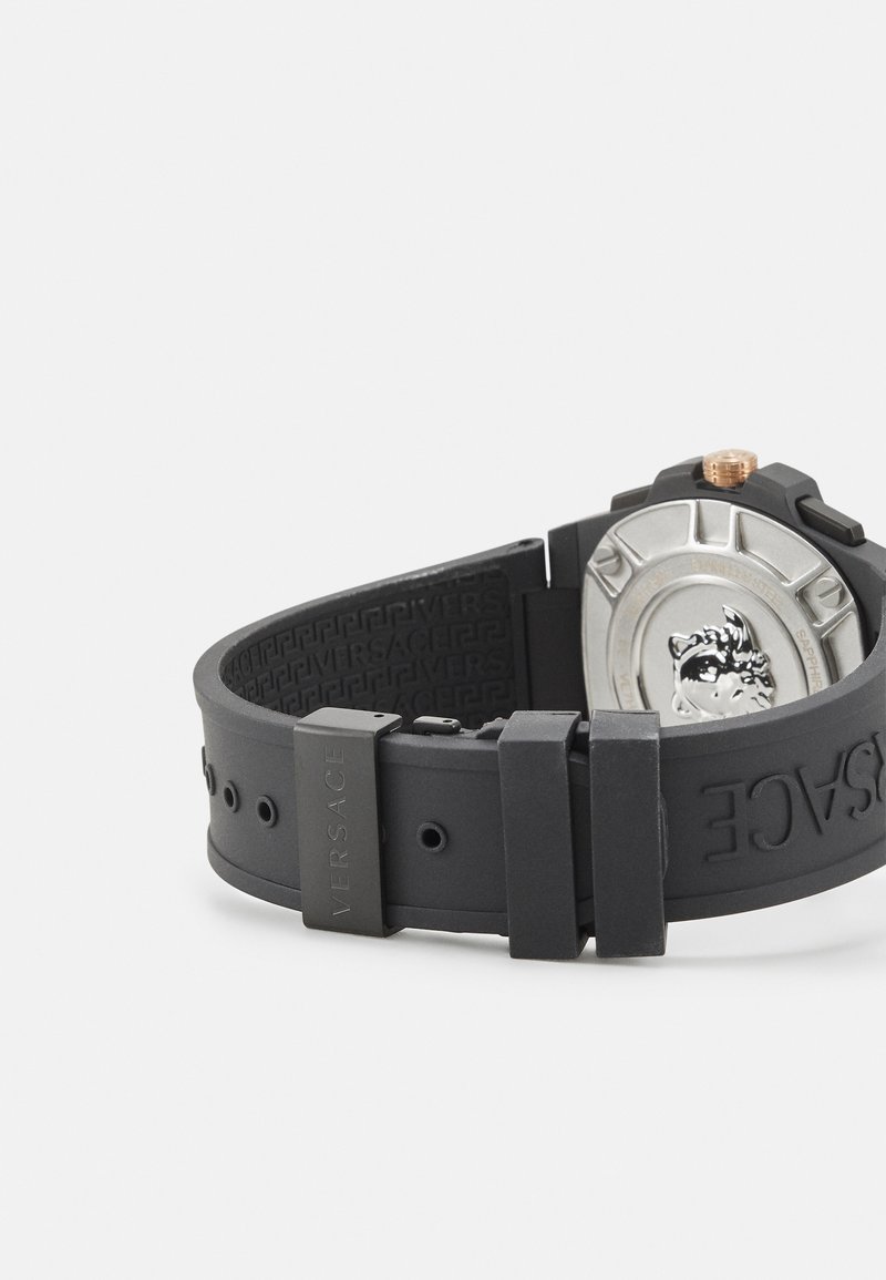 Svart gummiband för klocka med en urtavla i silver och guld, med upphöjda detaljer och en präglad logotyp på bandet.