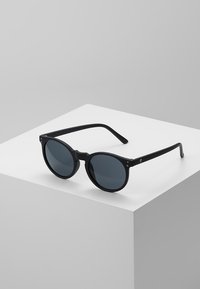 Ochelari de soare rotunzi, negri, cu lentile întunecate, finisaj mat elegant și brațe subțiri cu un detaliu discret al logo-ului. Așezați pe un fundal deschis.