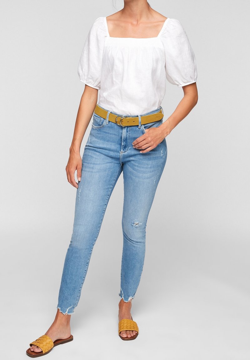 Femme portant un haut blanc à manches bouffantes, un jean skinny déchiré bleu clair, une ceinture jaune moutarde et des sandales tissées assorties, debout devant un fond neutre.