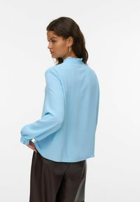 Una donna con capelli scuri indossa una blouse a maniche lunghe di colore azzurro chiaro con colletto e polsini arricciati, abbinata a pantaloni marrone scuro, in posa di profilo.